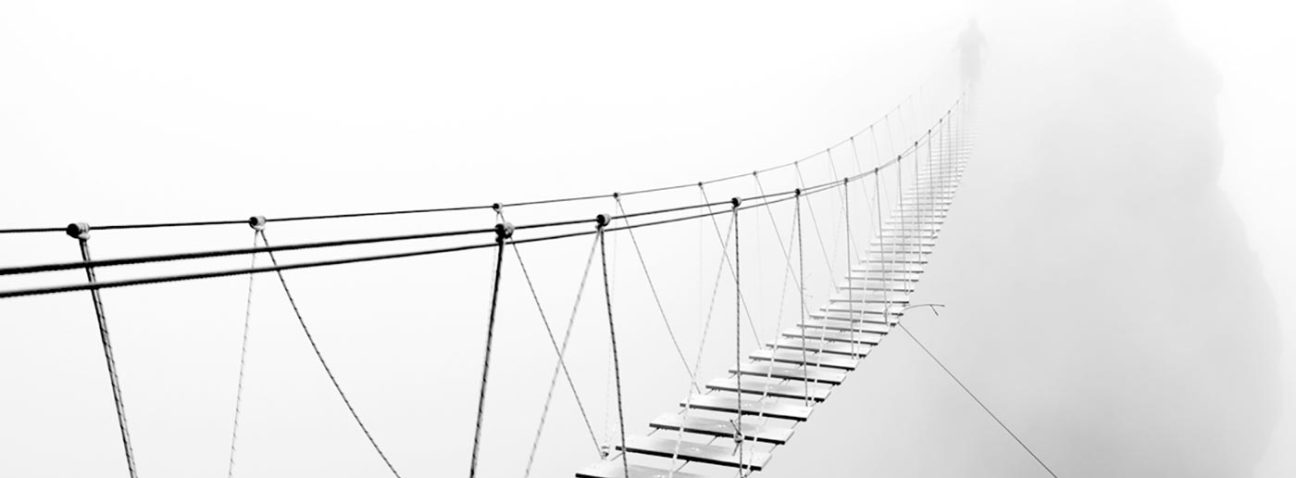 Skinny wooden bridge spanning a gap with a fiigure visible in the fog